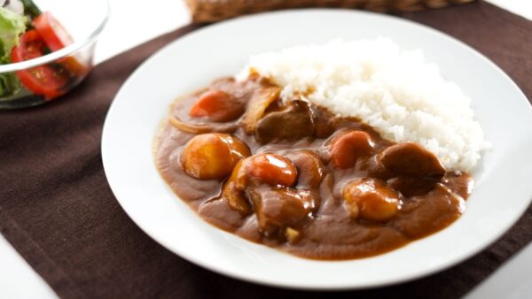 A plate of Japanese curry with rice, vegetables, and a side salad. The curry is a thick, brown sauce with chunks of meat and vegetables.