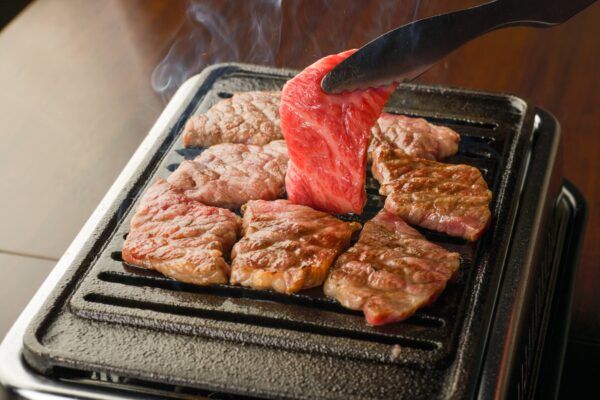 A plate of thinly sliced Halal A5 Wagyu beef being grilled on a yakiniku grill. The beef is sizzling and smoke is rising from the grill.