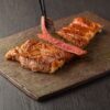 A perfectly cooked steak, sliced and served on a wooden board. Chopsticks are holding a piece of the steak. There is a small dish of wasabi and salt on the side.