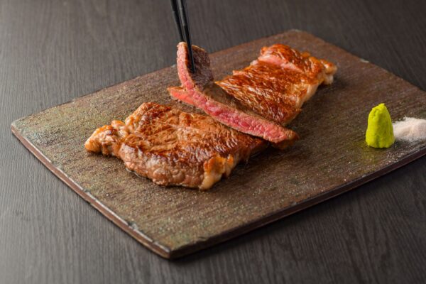 A perfectly cooked steak, sliced and served on a wooden board. Chopsticks are holding a piece of the steak. There is a small dish of wasabi and salt on the side.