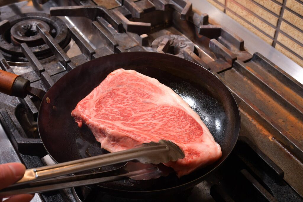 A thick, A5 marbled Wagyu steak sizzling in a cast iron skillet, being flipped with tongs.