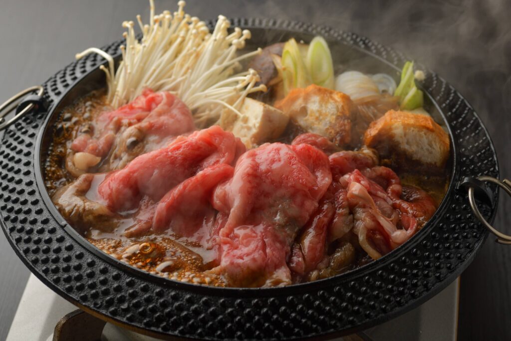 A sizzling hot pot filled with a dark broth, containing thinly sliced halal wagyu beef, enoki mushrooms, tofu, and other vegetables. Steam rises from the pot.