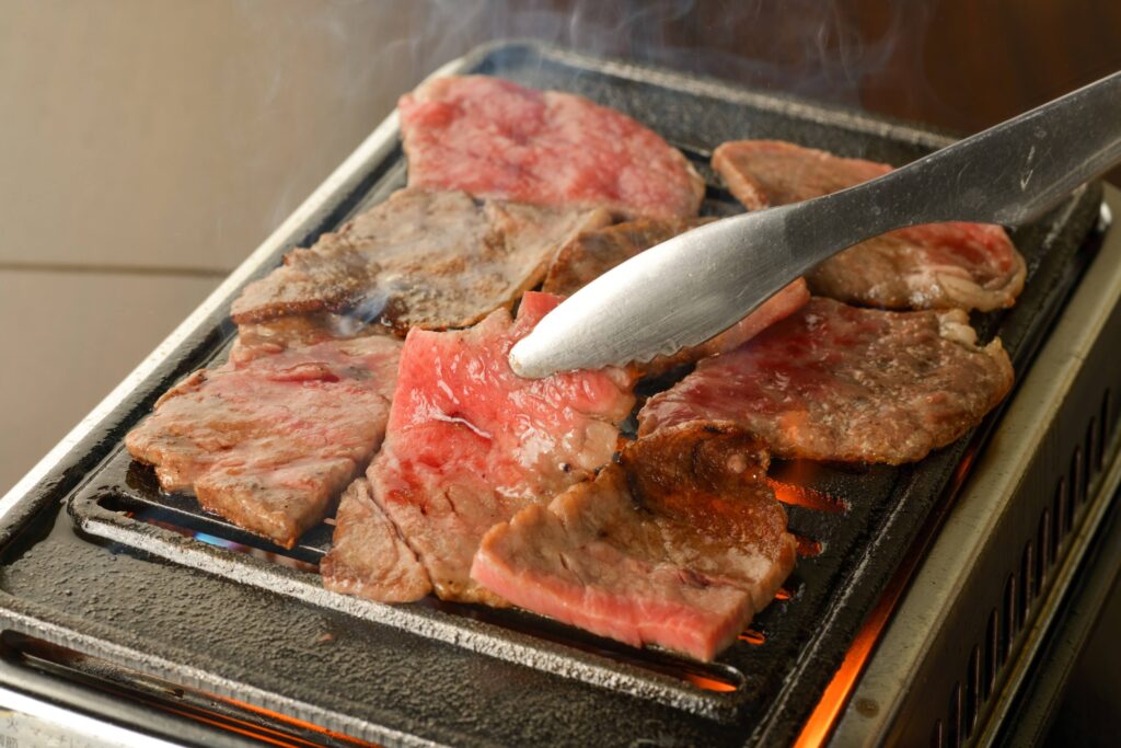 A close-up of thinly sliced halal wagyu beef being grilled on a hot plate. The wagyu beef is sizzling and has a reddish-pink color.